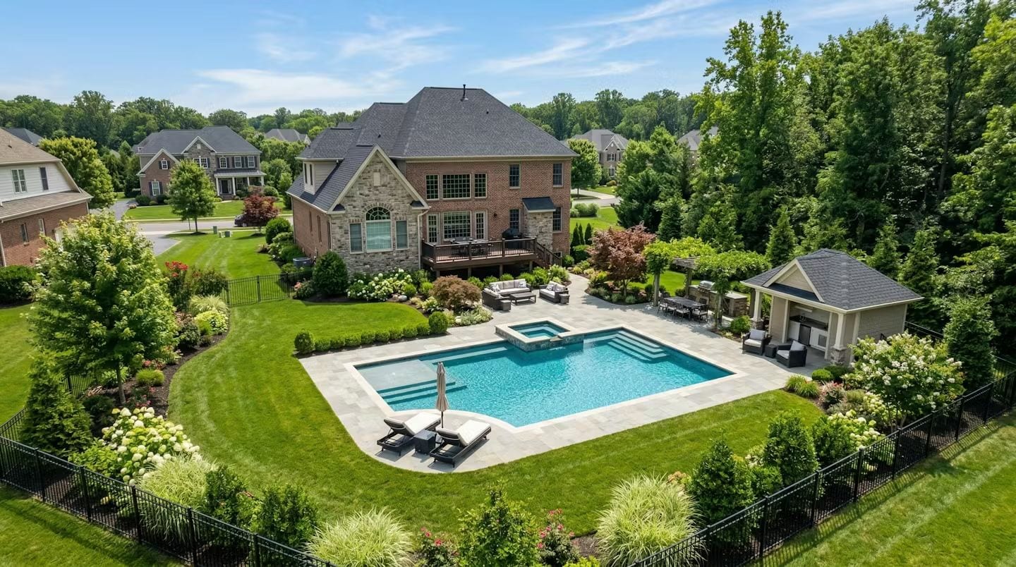 Aerial view of a luxury brick and stone colonial home in a Virginia or Maryland suburb with a rectangular inground pool and attached spa on a travertine patio, a pool house pavilion, outdoor seating area, and manicured gardens enclosed by a black iron fence surrounded by mature trees