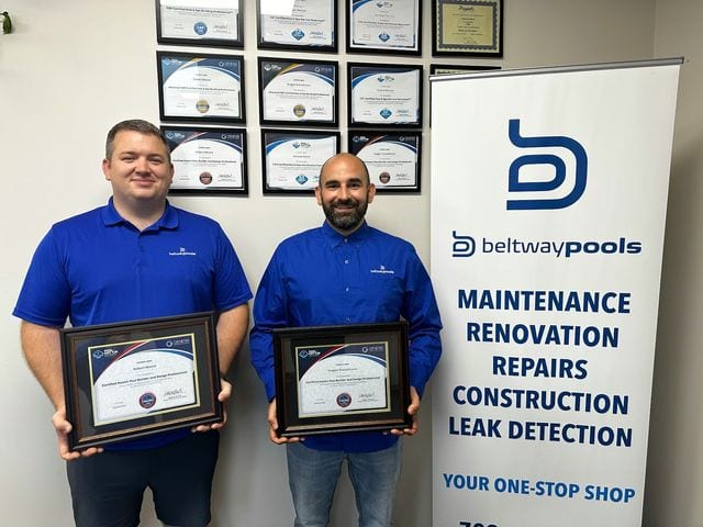 Dragan and Rob, Beltway Pools team members, in branded blue polo shirts holding their Genesis Certified Master Pool Builder and Design Professional certificates, standing in front of a wall displaying multiple PHTA and Genesis professional pool industry certifications, next to a Beltway Pools service banner listing maintenance, renovation, repairs, construction, and leak detection