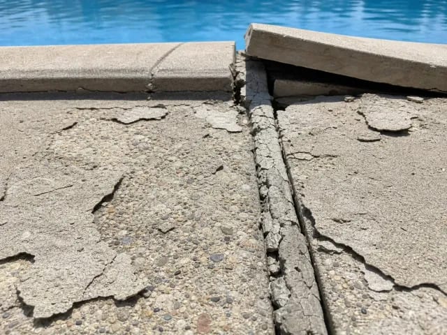 Spalled concrete pool deck with a cracked open expansion joint beside lifted stone coping