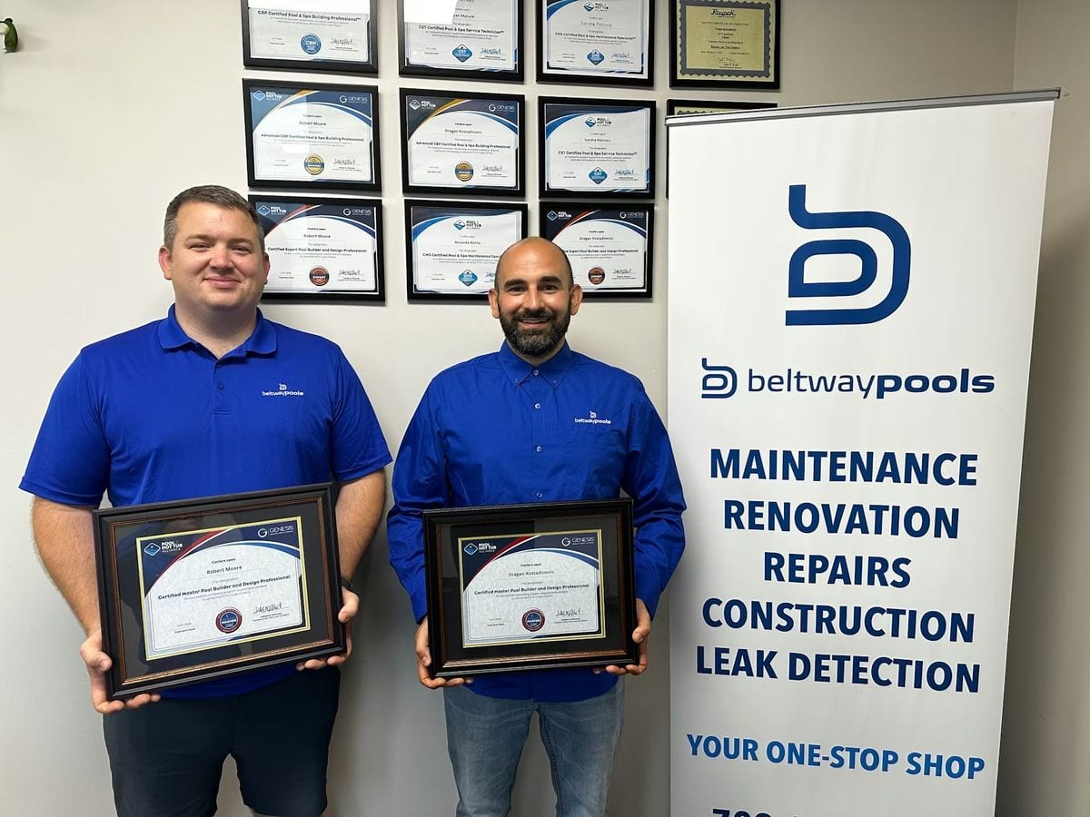 Dragan and Rob, Beltway Pools team members, in branded blue polo shirts holding their Genesis Certified Master Pool Builder and Design Professional certificates, standing in front of a wall displaying multiple PHTA and Genesis professional pool industry certifications, next to a Beltway Pools service banner listing maintenance, renovation, repairs, construction, and leak detection