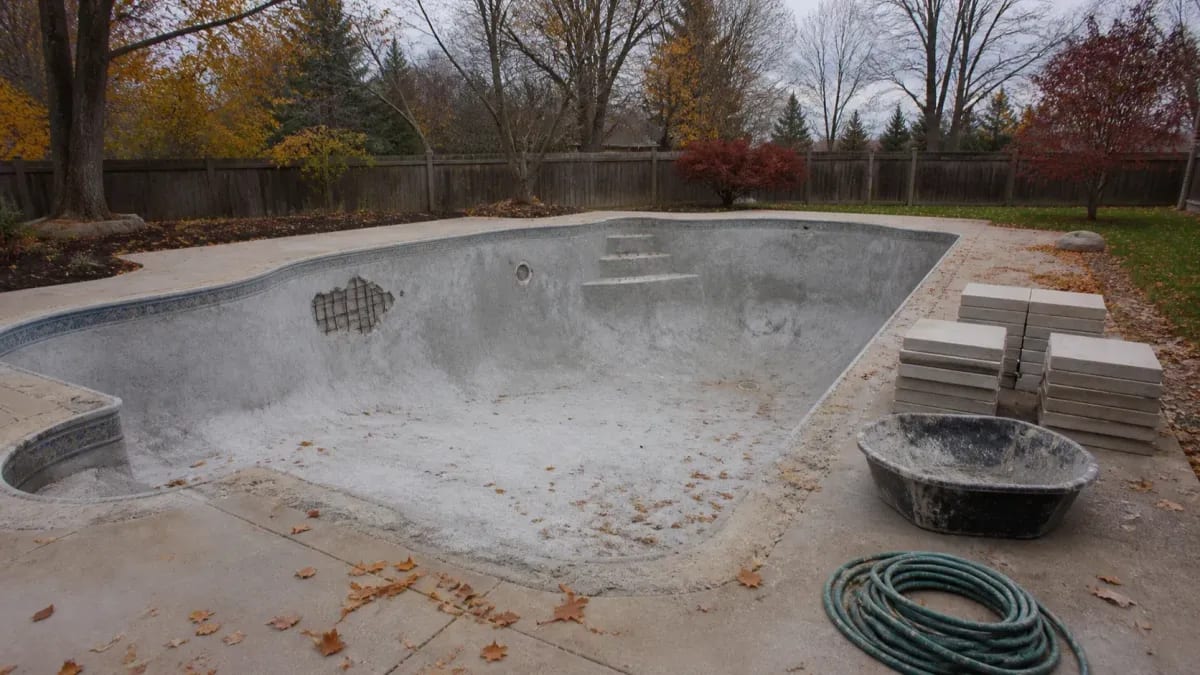 Drained gunite pool shell with an exposed rebar patch, stacked coping stones, and autumn leaves on the deck
