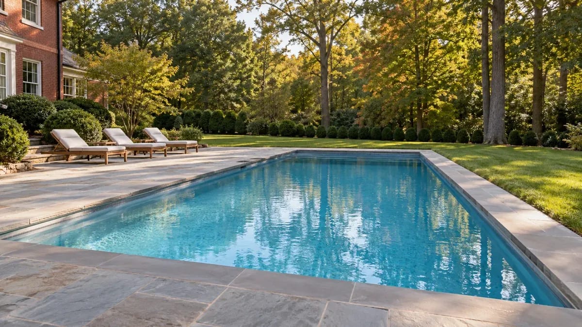 Rectangular inground pool with stone coping beside a brick house, with lounge chairs, lawn, and early-fall trees.