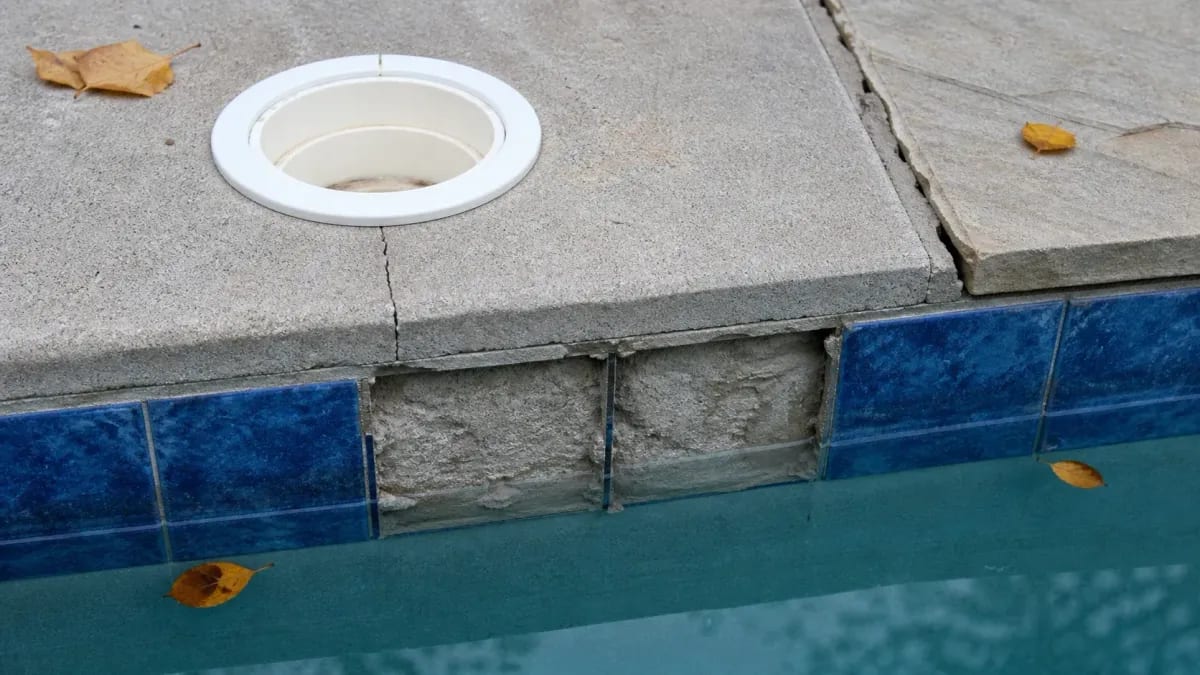 Cracked pool deck beside a round skimmer lid, two waterline tiles missing above the water, fallen autumn leaves